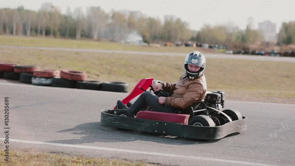 Man in a kart with a crash helmet on his head waiting looking back, starts moving and stops. Side view. Go-kart track.