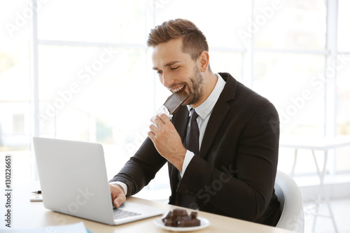 Young man eating chocolate while working with laptop in office