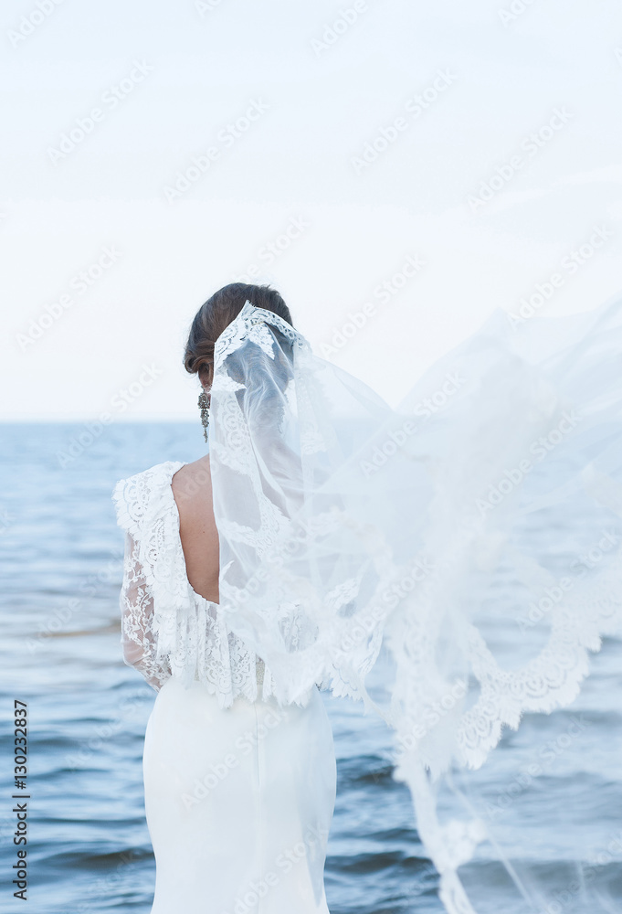 Naklejka premium The bride in a beautiful dress and vintage faie looking at the sea. Dress with open back. veil flies. Wedding dress with lace. Wedding photo concept. Bride. Art wedding photo