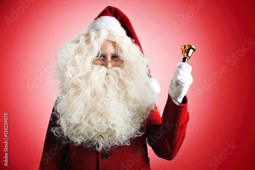 Santa Claus with huge pretty beard in red hat and coat ringing a golden bell on red background photo