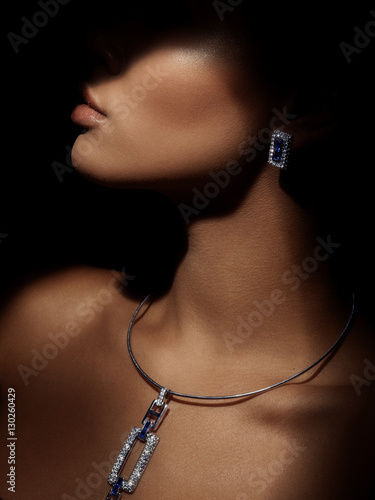 Portrait of an elegant and beautiful young smartly dressed woman with sparkling jewelry made from precious metals on her neck. Bright sapphire earrings and gorgeous necklace with diamonds. Darkness. photo