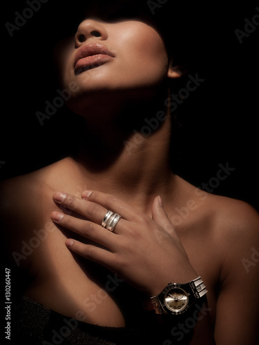Portrait of an elegant, gorgeous and mystery woman with luxury wide rings made from precious metals on her finger and watch on your wrist. Dark studio background. Shadow on the face. Elegance.