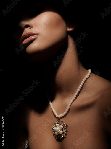 Portrait of an elegant and mystery smart lady with luxury jewelry made from precious metals on her neck (gorgeous silver necklace with pearls). Dark studio background. Shadow on the face.