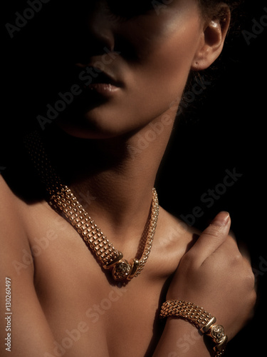 Portrait of an elegant and beautiful young smartly dressed woman with luxury jewelry made from precious metals on her neck (gorgeous necklace with diamonds). Golden bracelet is on her wrist. Shadow.