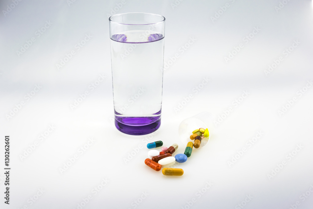 Many colored tablets and capsules near a glass of water with purple bottom isolated on white background. Many colored tablets and capsules near a glass of water with purple bottom isolated on white background.