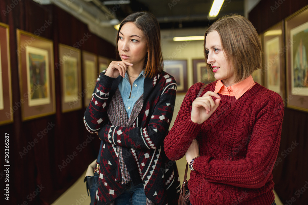 Two girl friends standing in a gallery contemplating and discuss paintings artworks displayed on gallery walls Two girl friends standing in a gallery contemplating and discuss paintings artworks displayed on gallery walls