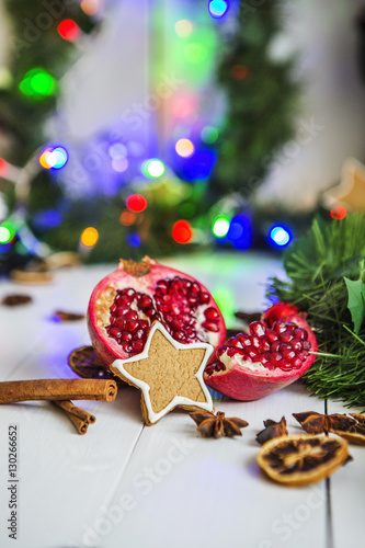 Имбирный пряник в виде звезды, разрезанный красный гранат, корица, сушеные лимоны лежат на белом деревянном столе на фоне зеленой новогодней гирлянды и новогодних огней.
