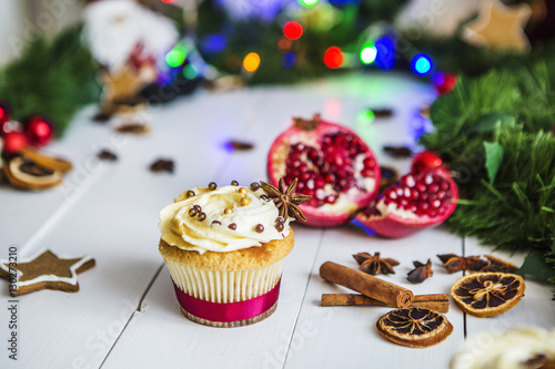 Пирожное с кремом, капкейк, мбирный пряник в виде звезды, разрезанный красный гранат, корица, сушеные лимоны лежат на белом деревянном столе на фоне зеленой новогодней гирлянды и новогодних огней.