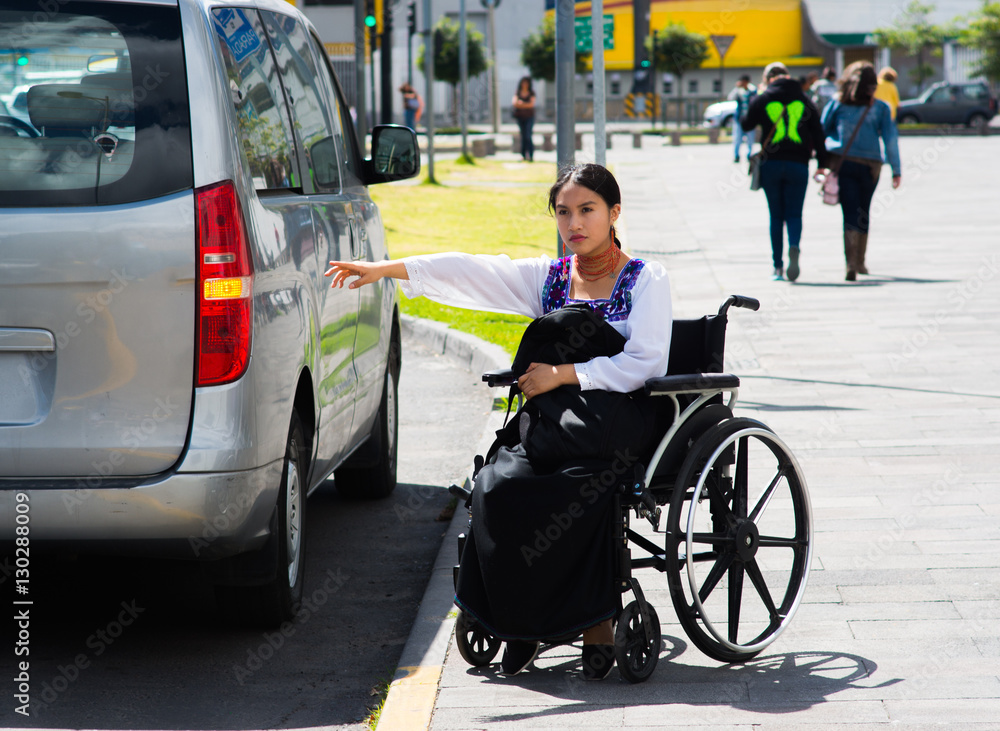 Obraz premium Young brunette woman sitting in wheelchair smiling with positive attitude, holding out arm looking for taxi, outdoors environment, physical recovery concept