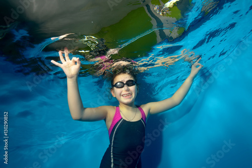Sports little girl swims underwater in the pool on a blue background, looking at camera and smiling. Portrait. Shooting underwater. Landscape orientation