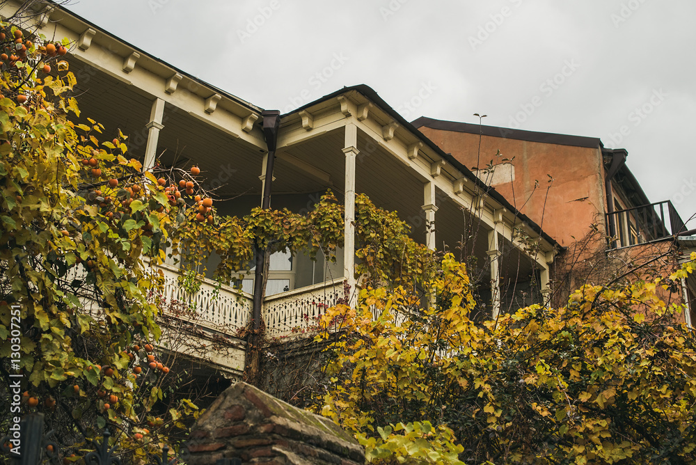 Typical house exterior of Tbilisi old town center in Autumn. Shabby house with wooden balcony, fruit trees and twining shrubs Typical house exterior of Tbilisi old town center in Autumn. Shabby house with wooden balcony, fruit trees and twining shrubs