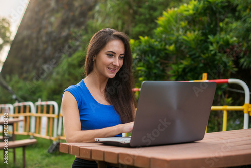 Young pretty smiling student chatting with friends on laptop in the summer park.