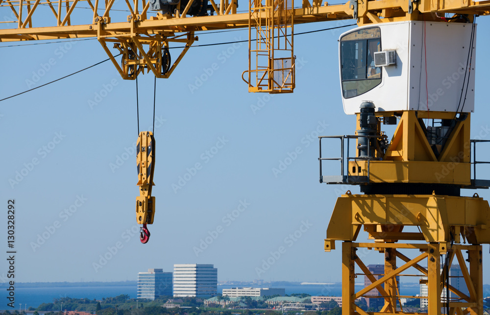 Huge yellow building construction crane with hook, pulley, lattice boom and cabin with an aerial view of a city and blue water bay.