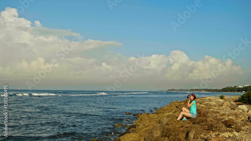 Young woman with red hair in glasses photographs sea seating on the rocks, beach of Dominican Republic