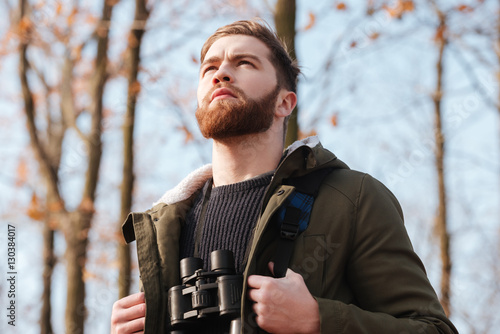 Serious bearded young man with field-glass standing in forest