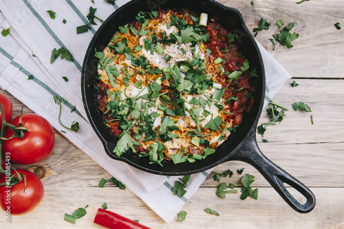 Delicious fresh morning Shakshuka on the wooden rustic table with some tomatoes on the side photo