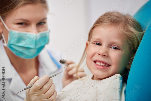 Happy smiling child sitting on dental chair ready to cure teeth. Portrait of woman dentist in mask with tools treating patient. 