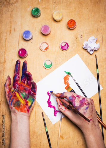 Hands of the artist in bright colored paint with brush on wooden table. Woman draws on a sheet of paper