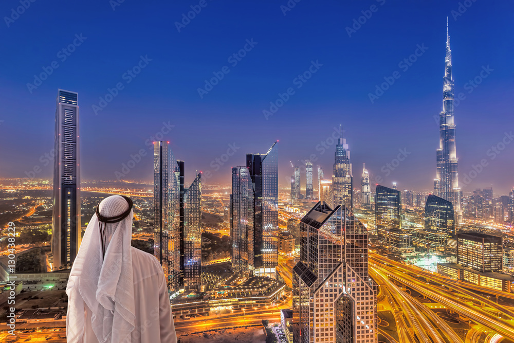 Obraz premium Arabski mężczyzna ogląda nocny panoramę Dubaju z nowoczesną futurystyczną architekturą w Zjednoczonych Emiratach Arabskich
