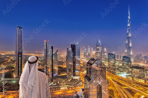 Photographie Arabian man watching night cityscape of Dubai with modern futuristic architectur