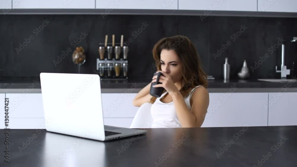 Sad bored young woman using laptop and drinking coffee while sitting on a kitchen Sad bored young woman using laptop and drinking coffee while sitting on a kitchen