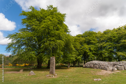 Inverness, Scotland - June 2, 2012: Wide shot of the menhir stone circle and grave site heap of gray stones at prehistoric Clava Cairns. Surrounded by green trees, grassy field.
