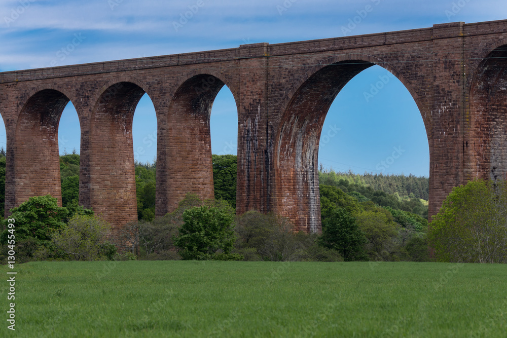 Inverness, Scotland - June 2, 2012: Section of red stone Culloden train viaduct with widest bow over Nairm River. Green meadows up front, blue sky, forests behind bridge. Water discoloring of walls. Inverness, Scotland - June 2, 2012: Section of red stone Culloden train viaduct with widest bow over Nairm River. Green meadows up front, blue sky, forests behind bridge. Water discoloring of walls.