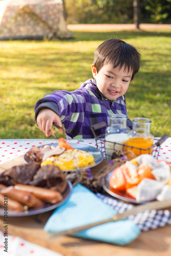 An Asian cute young boy eating breakfast outdoor at camping site in nature sunlight with a tent at the background.