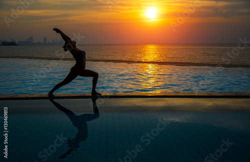 Silhouette young woman practicing yoga on swimming pool and the beach at sunset.