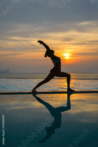 Silhouette young woman practicing yoga on swimming pool and the beach at sunset.