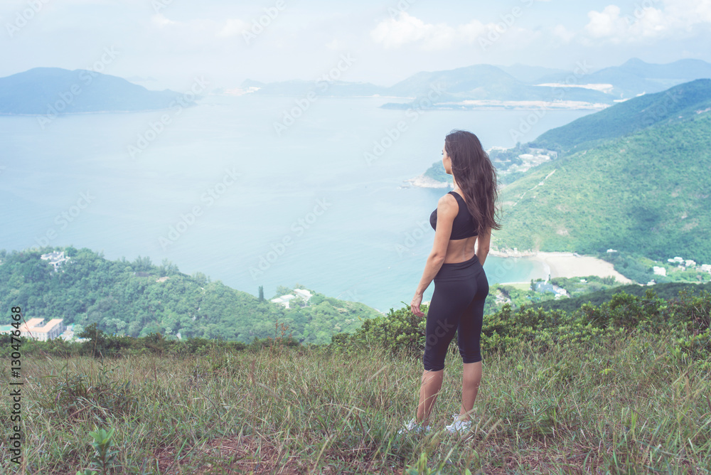Sporty young woman standing on hill admiring the view of sea and green mountains in sunlight