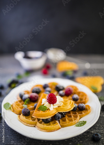 Homemade Belgian waffles with fresh ripe berries served on white plate and dark stone background.