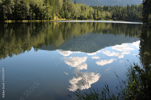 In the clear water of a forest lake reflects the sky, mountain, forest and clouds. Photo partially tinted. 