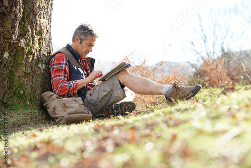 Hiker relaxing by tree looking at map and using tablet