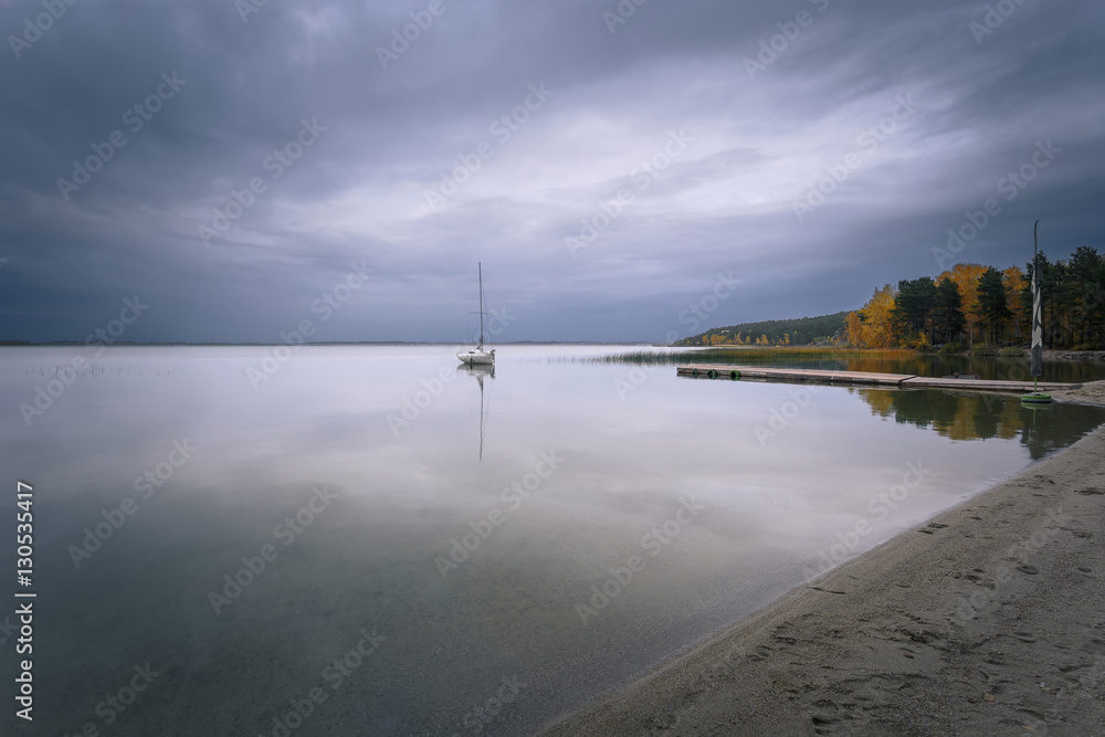 Autumnal cloudy landscape. A beautiful view at the lake with a lonely white yacht, moores near wooden pier.