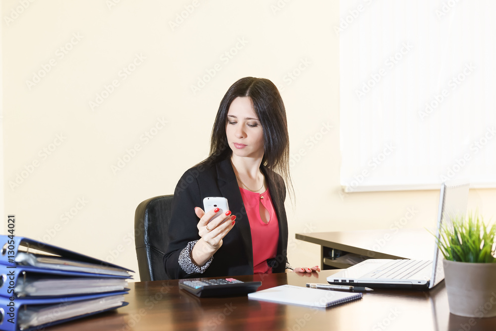 Young attractive woman in a business suit looks at the screen of the smartphone at the Desk in the office Young attractive woman in a business suit looks at the screen of the smartphone at the Desk in the office