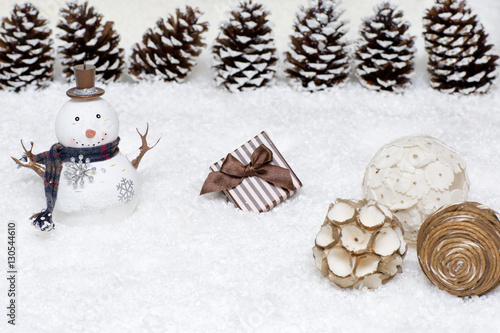 Gift box and snowman are in the snow. Christmas light background has cones. 