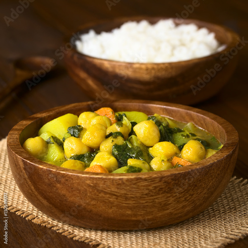 Veganischer Kichererbsen Curry mit Spinat, Kartoffeln und Karotten in Holzschüssel mit Reis im Hintergrund, fotografiert mit natürlichem Licht (Selektiver Fokus, Fokus ein Drittel in den Curry) photo