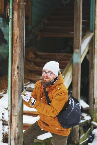 Young attractive smiling bearded man with glasses, next to the old dilapidated wooden staircase in the house on a cold winter day.