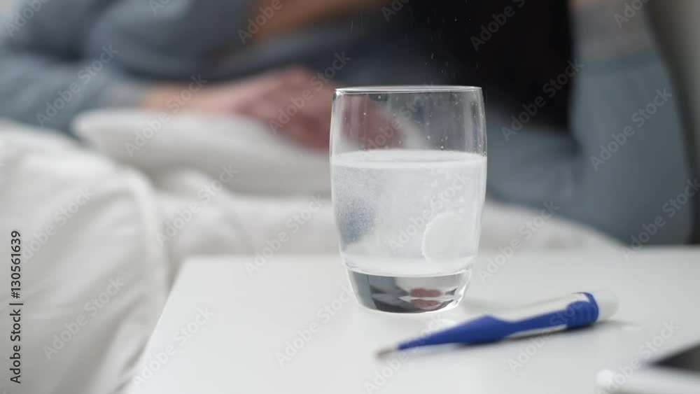 Painkiller tablet fizzing and dissolving into a glass of water and sick woman lying in bed on the background Painkiller tablet fizzing and dissolving into a glass of water and sick woman lying in bed on the background