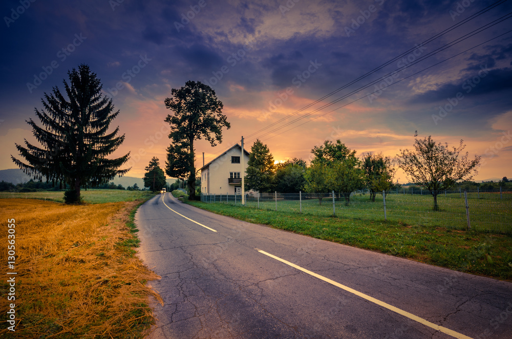 Road beside wheat field and cottage at sunset with colorful overcast sky.