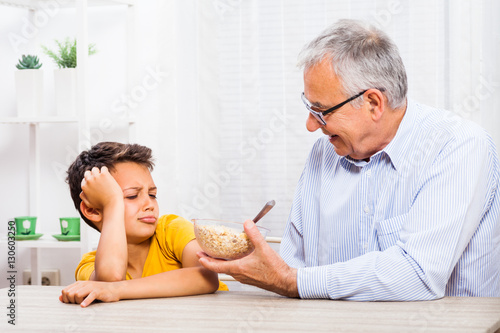Grandfather is giving oatmeal to his grandson but he refuses to eat it. 