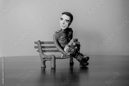 Plasticine figure of man with a bouquet of red flowers in hands, sitting on a bench made of wood