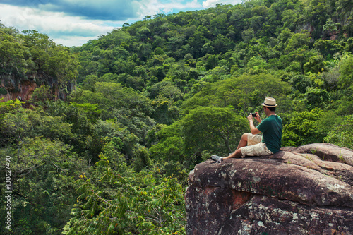 Young man tourist sittig on the rock at the top of mountain and taking photo with smartphone of a green tropical valley. 25-30 years old with t-shirt, shorts and hat.
