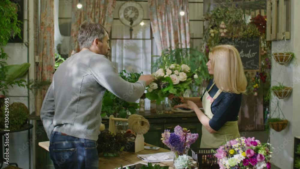 Beautiful mid-adult florist showing man bouquet of white roses and telling him something with smile in flower shop Beautiful mid-adult florist showing man bouquet of white roses and telling him something with smile in flower shop