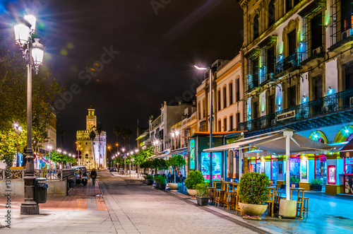 night view of a lively street in the city center of sevilla with the illuminated torre de oro at ist end