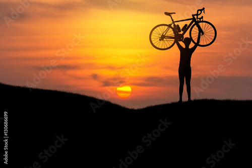 Silhouette the man stand in action lifting bicycle above his head on the meadow with sunset 