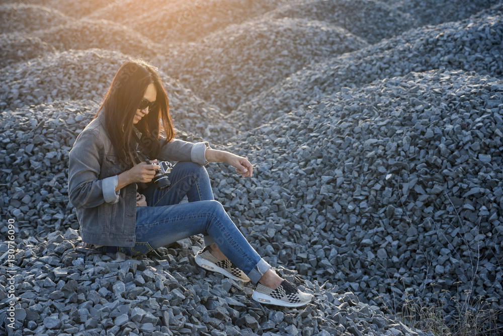 A beautiful woman wearing glasses, jacket and blue jeans,holding camera sitting on a pile of rocks
