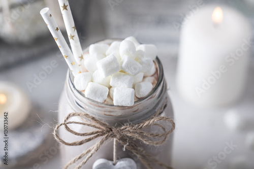 Cocoa with marshmallow and straws in the glass jar decorated with little white heart on the table with candles for the winter holidays. Close up