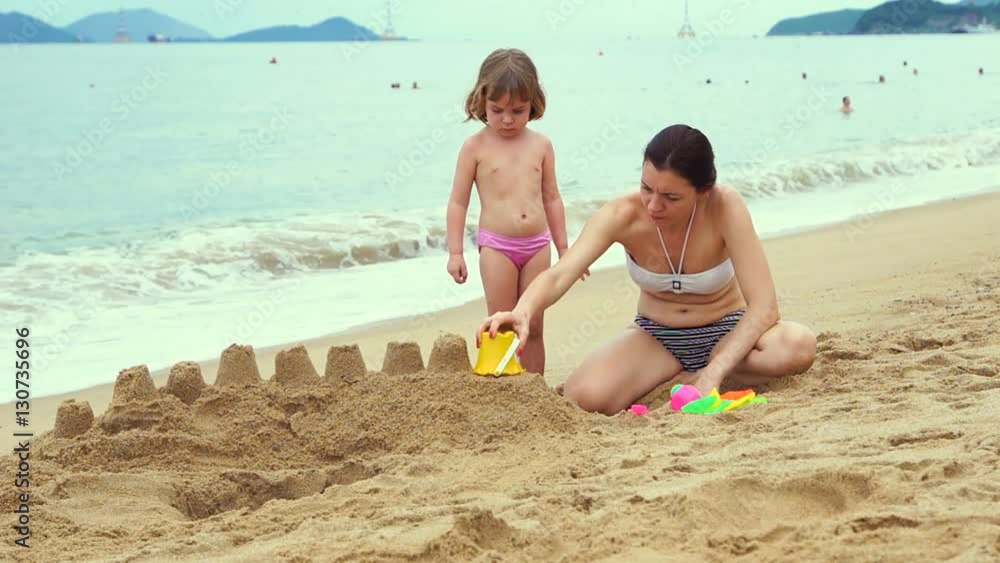 Attractive mother and small child building a sand castle. Sea shore. Unrecognizable people on the background. Attractive mother and small child building a sand castle. Sea shore. Unrecognizable people on the background.